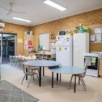 Spacious Y WA OSHC room with round tables, chairs, kitchen area, and open door to outdoor space.