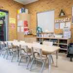 Y WA OSHC room with long activity table, chairs, craft storage shelving, and open doors to outdoor area.