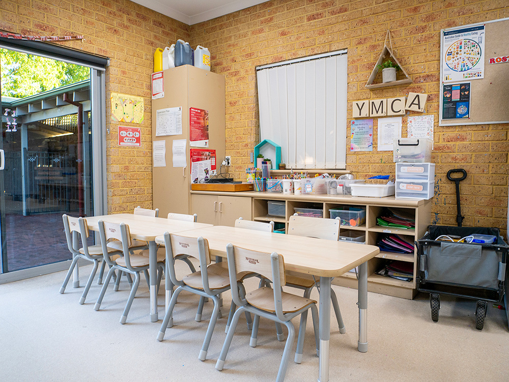 Y WA OSHC room with long activity table, chairs, craft storage shelving, and open doors to outdoor area.