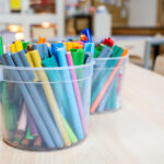 Two clear buckets filled with colourful markers and felt-tip pens on a table in an OSHC craft area.