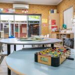 OSHC room with round tables, foosball game, books, storage shelving, and sliding doors to outdoor area.