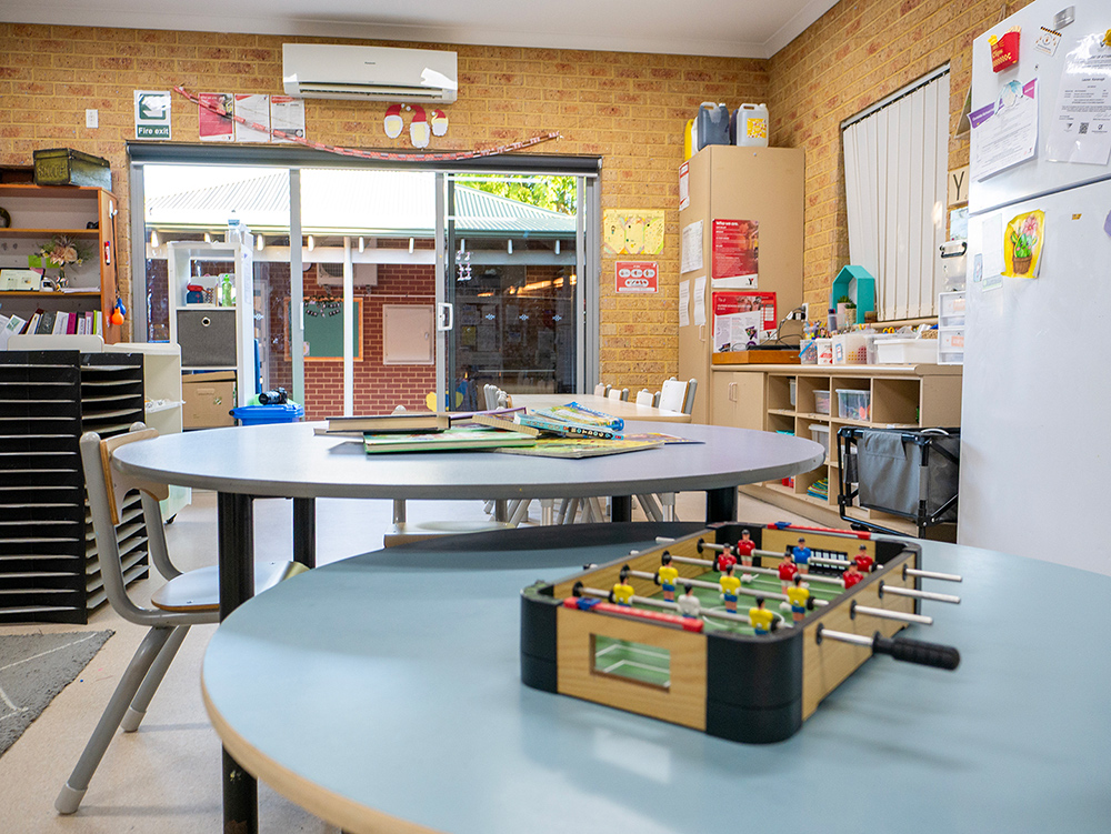 OSHC room with round tables, foosball game, books, storage shelving, and sliding doors to outdoor area.