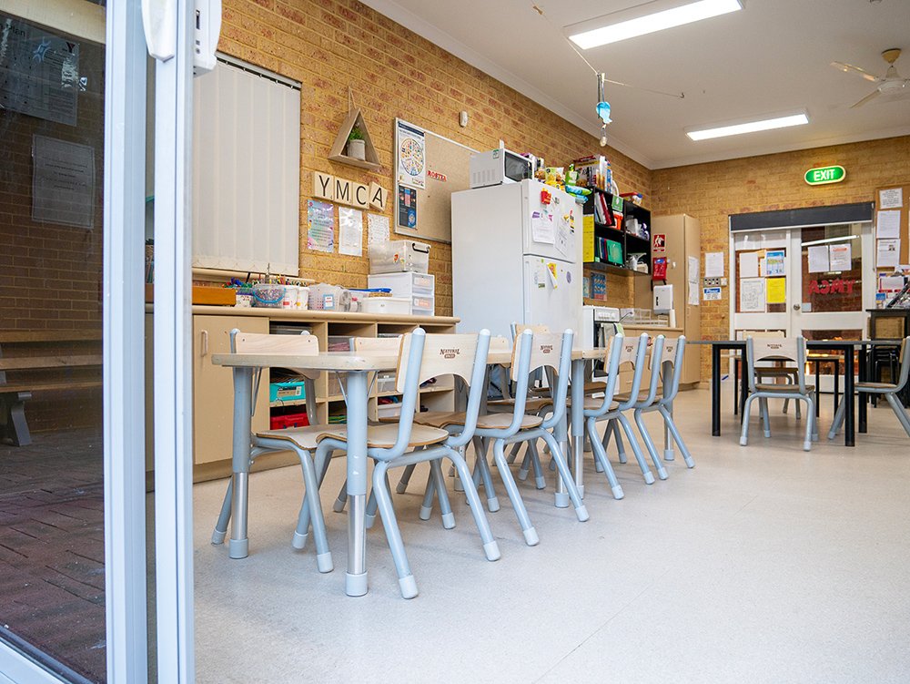 Y WA OSHC activity room with long tables, chairs, storage shelving, fridge, and brick feature wall.