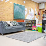 Inviting OSHC room with grey sofa, chalkboard, bookshelf, activity tables, and children's artwork on brick walls.