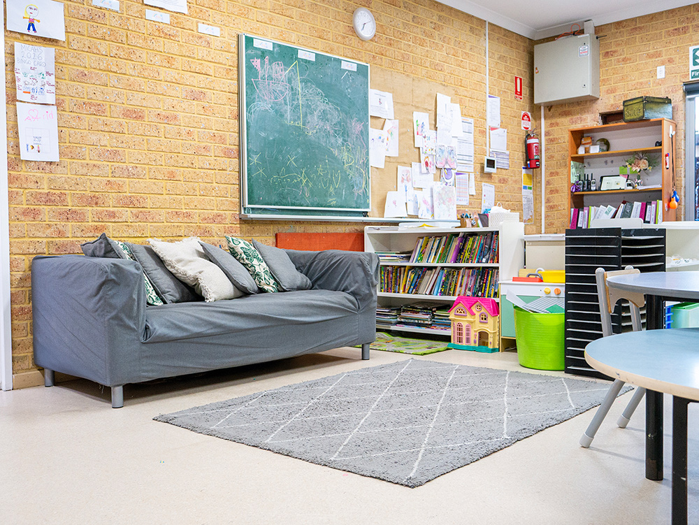 Inviting OSHC room with grey sofa, chalkboard, bookshelf, activity tables, and children's artwork on brick walls.