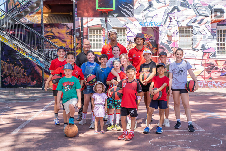 Children and Perth Wildcats players pose together on an outdoor basketball court with colourful murals.