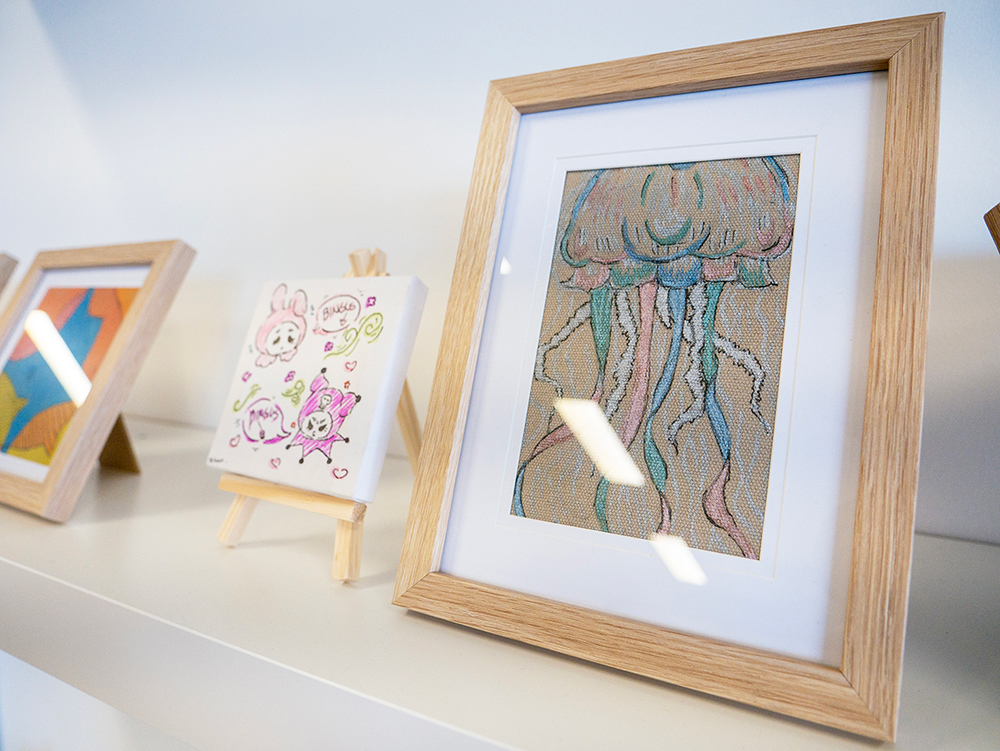 A shelf displaying framed artworks created by young people, including a detailed jellyfish drawing and colourful character illustrations on mini easels