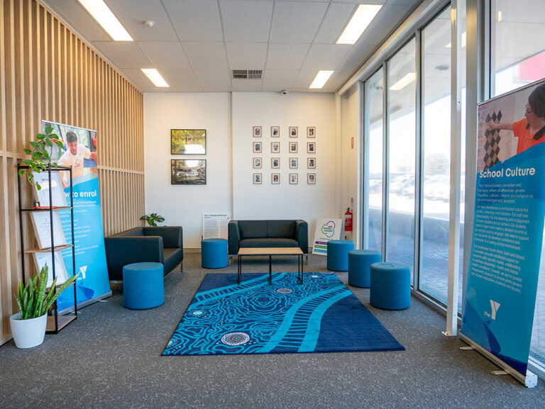 A welcoming Y School reception area featuring a seating area with an Aboriginal-designed rug, framed photos on the wall, indoor plants, and Y School informational banners