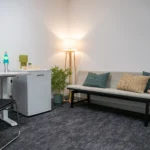 A cosy corner at the Y School with a couch, table, warm lighting and plants. There is also a lava lamp on the desk