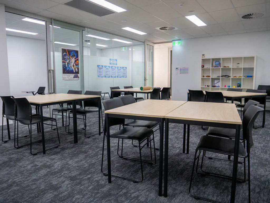 Large classroom at Y School Mirrabooka Junior Campus, featuring four tables and chares with a bookshelf in the back and artwork on the walls