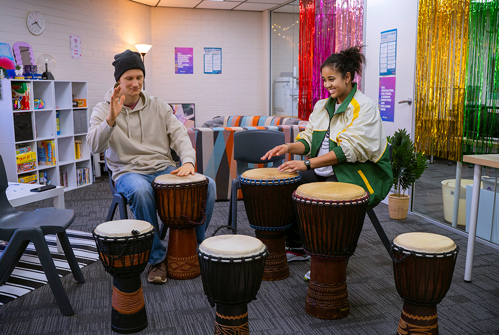 Two young people smiling and playing djembe drums together in a colourful youth centre room decorated with rainbow tinsel curtains