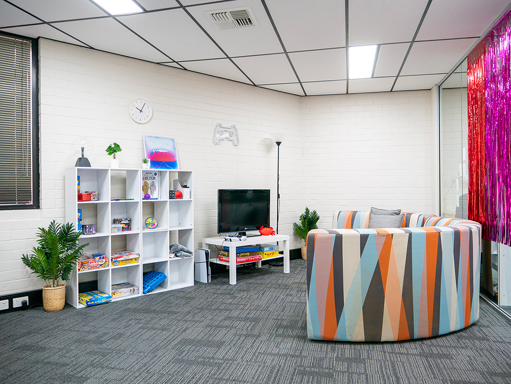 A bright and welcoming room featuring a colourful geometric circular sofa, gaming setup, shelving filled with toys and games, and a vibrant pink tinsel curtain