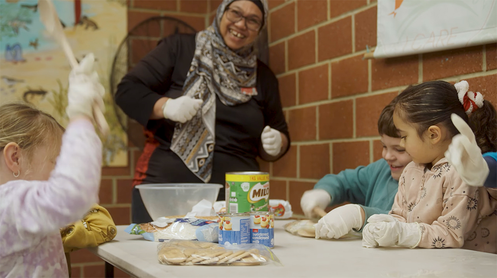 A smiling Y WA staff member wearing gloves supervises children doing a cooking activity at a table.