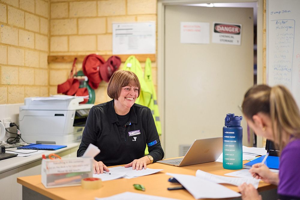 Two Y WA staff members smiling and working together at a desk with a laptop and paperwork.