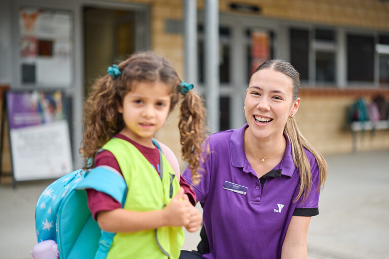 A smiling Y WA staff member in a purple polo crouches beside a young child with a backpack outside a school.