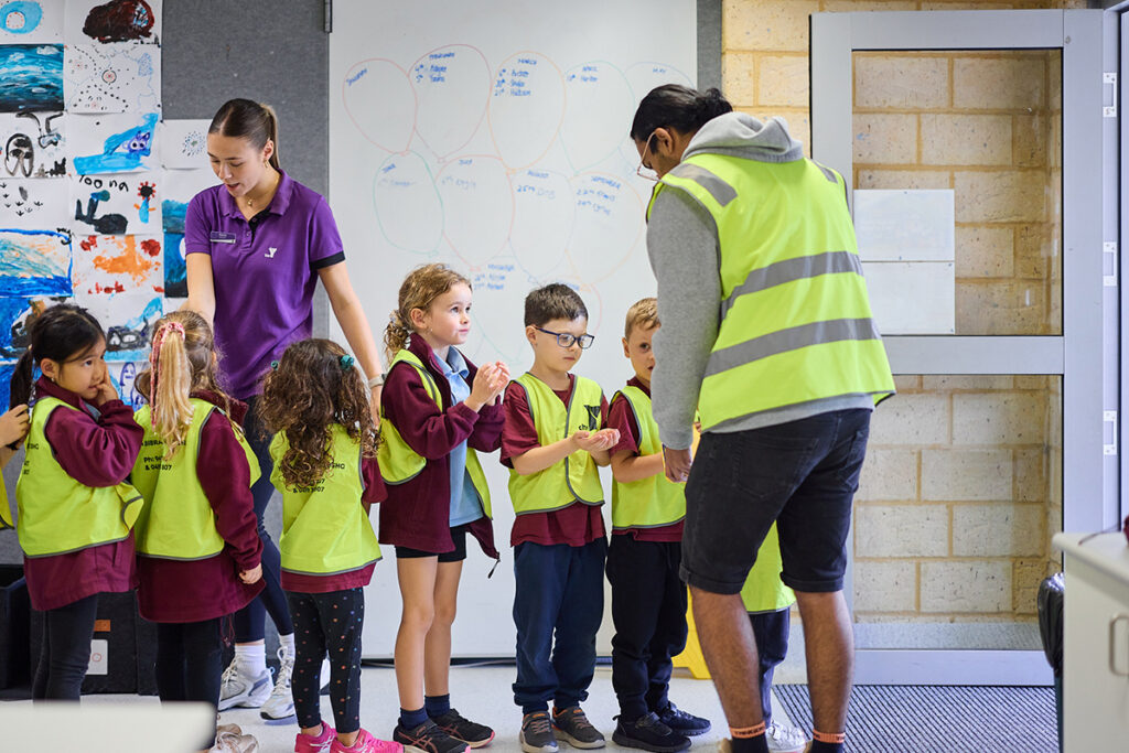 Y WA staff in purple and hi-vis vests supervise a group of young children in hi-vis vests inside a school room.