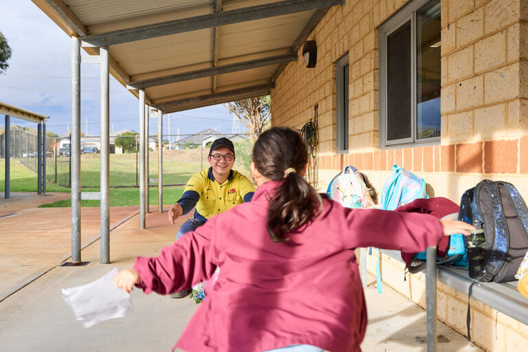 A smiling man in a hi-vis yellow shirt crouches to greet a child running toward him with open arms in a school verandah area