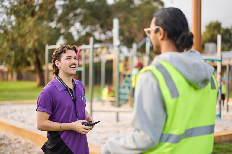 A man in a purple polo shirt laughs while talking with a person in a hi-vis vest on a school playground