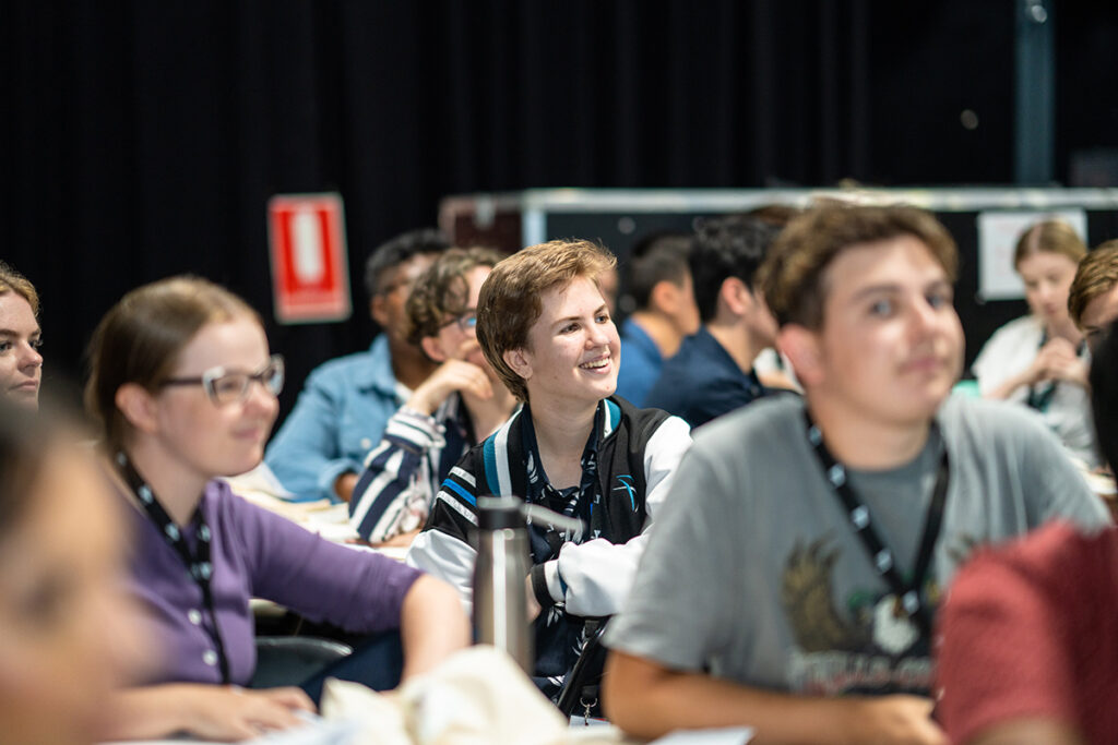 A smiling young woman listens attentively among a group of young people seated at tables during a conference or forum