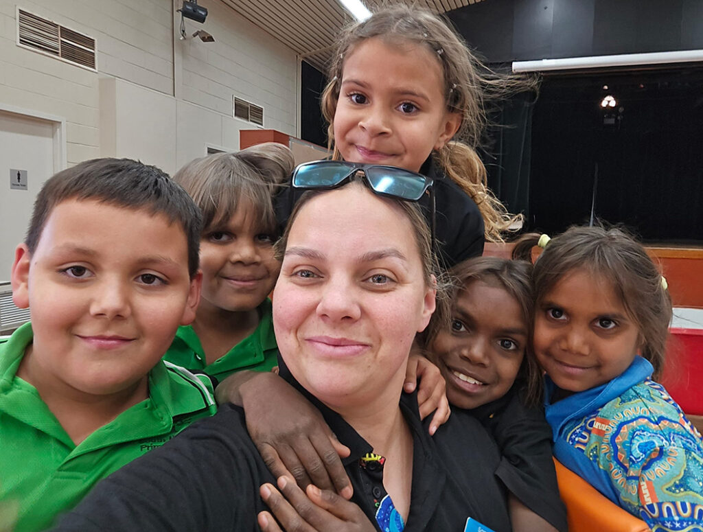 A youth worker takes a selfie with five smiling children crowded around her inside a community hall