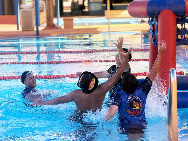 Three young people laugh and leap for the ball beside an inflatable basketball hoop during a pool game