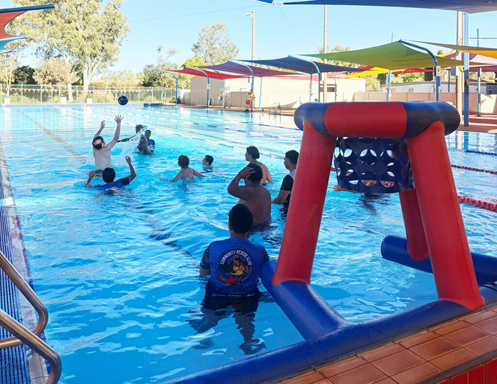 A group of young people play water basketball in an outdoor pool using an inflatable hoop on a sunny day