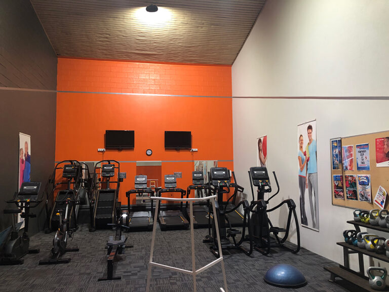 A gym cardio area with treadmills, ellipticals, and exercise bikes against an orange feature wall with two wall-mounted TVs