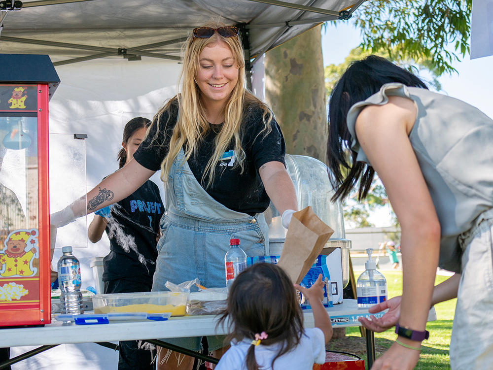 A Y WA youth worker happily passing a bag of popcorn to a parent and child at an event
