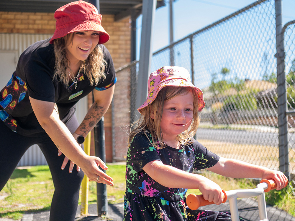 Educator pushing child on a bike in playground of Timber Tots childcare centre