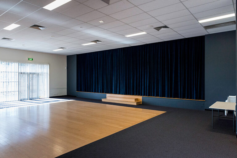 An empty multipurpose room with a timber floor, dark blue stage curtain, small stage, and natural light from a window