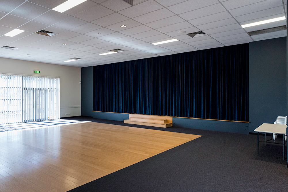 An empty multipurpose room with a timber floor, dark blue stage curtain, small stage, and natural light from a window