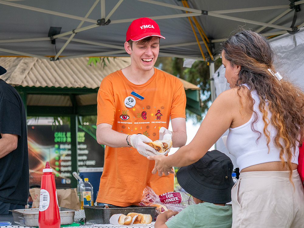 A Y WA youth worker happily passing a hot dog to a parent and child at an event