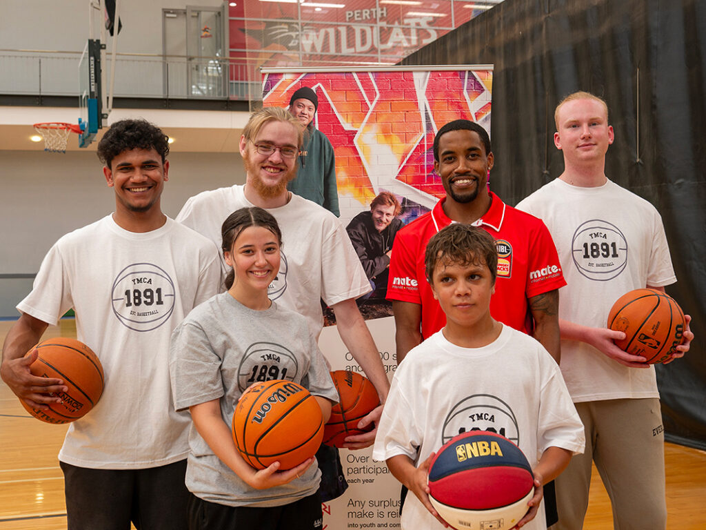 A group of young people in YMCA t-shirts hold basketballs alongside a Perth Wildcats player Bryce Cotton inside a basketball stadium