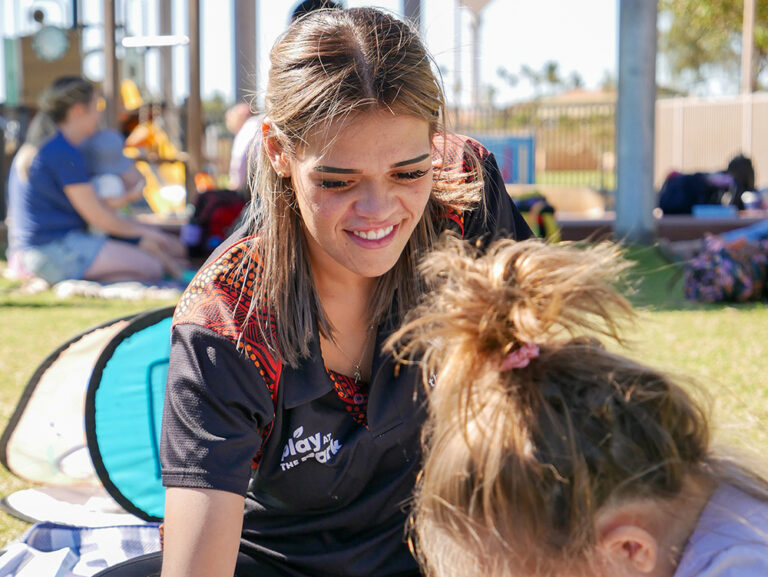 A smiling Y WA staff member in an Indigenous-print polo shirt engaging with a child outdoors on the grass.