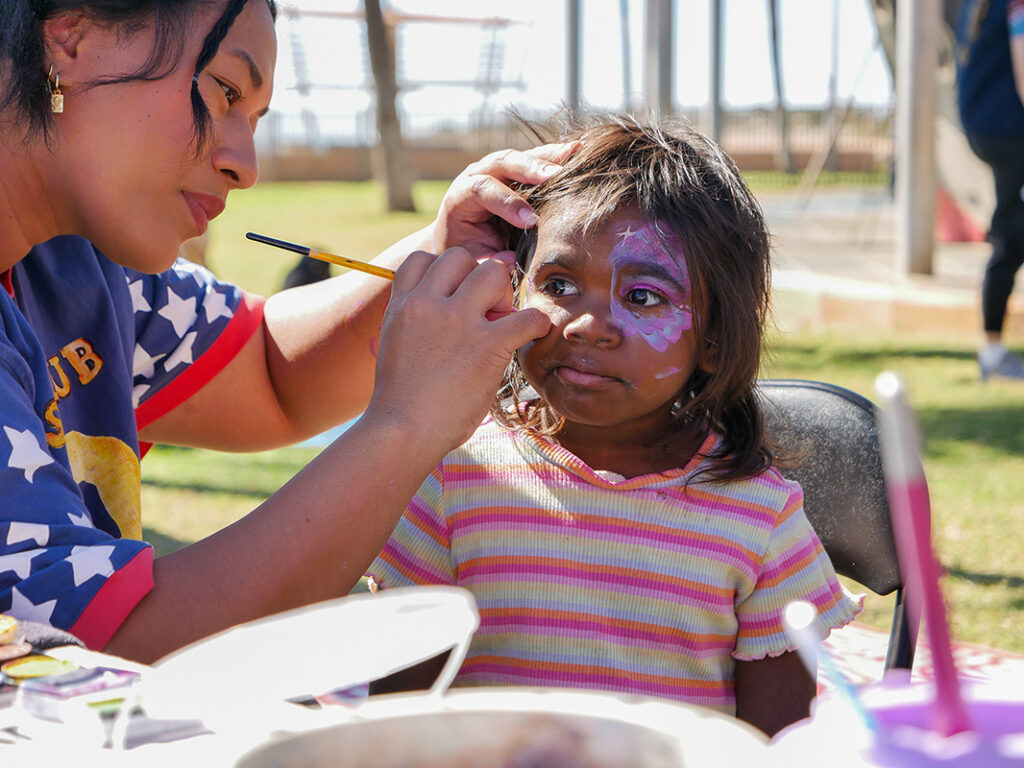 A woman carefully applies purple and white face paint to a young girl sitting outdoors at a community event