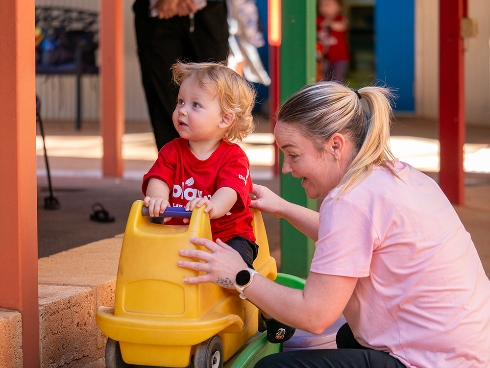A toddler in a red t-shirt sits on a yellow ride-on toy car while a smiling woman in a pink top crouches beside them in an outdoor play area