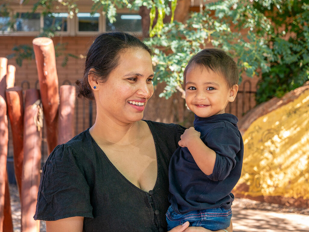 A smiling woman holds a toddler who grins at the camera in a sunny outdoor playground setting