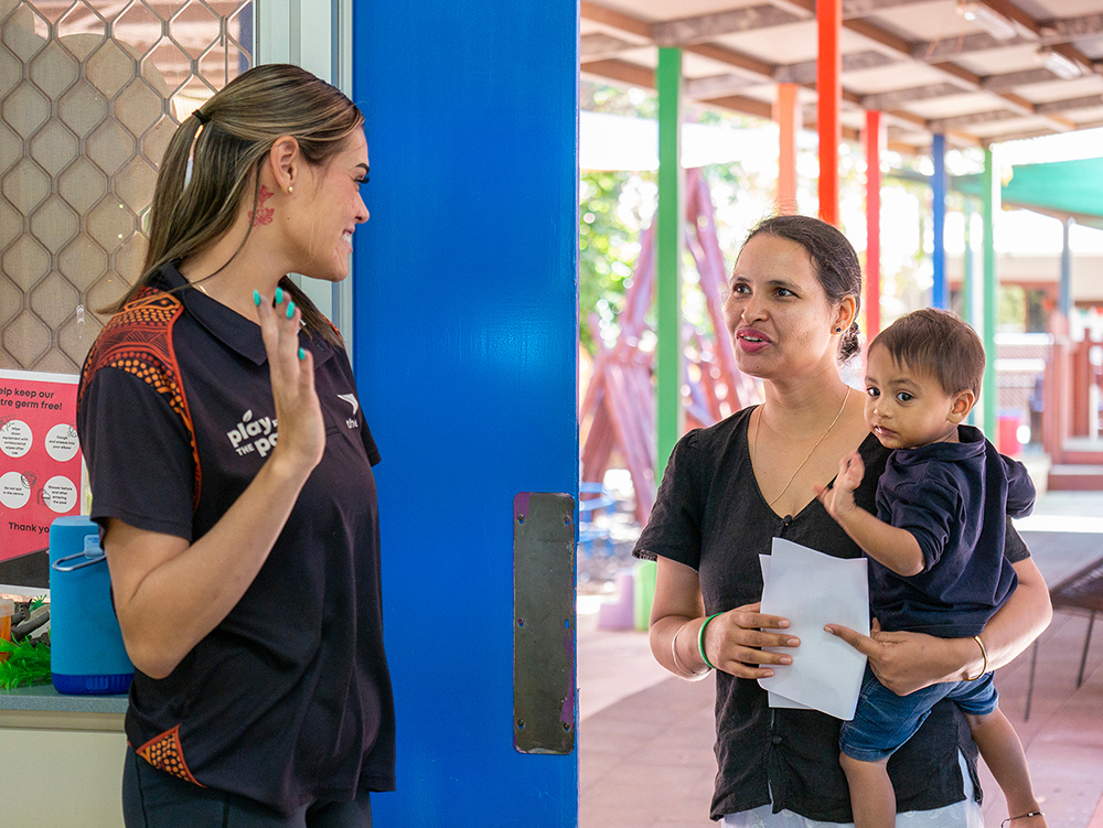 A childcare worker in a YMCA uniform smiles and chats with a woman holding a toddler at the entrance to a children's centre