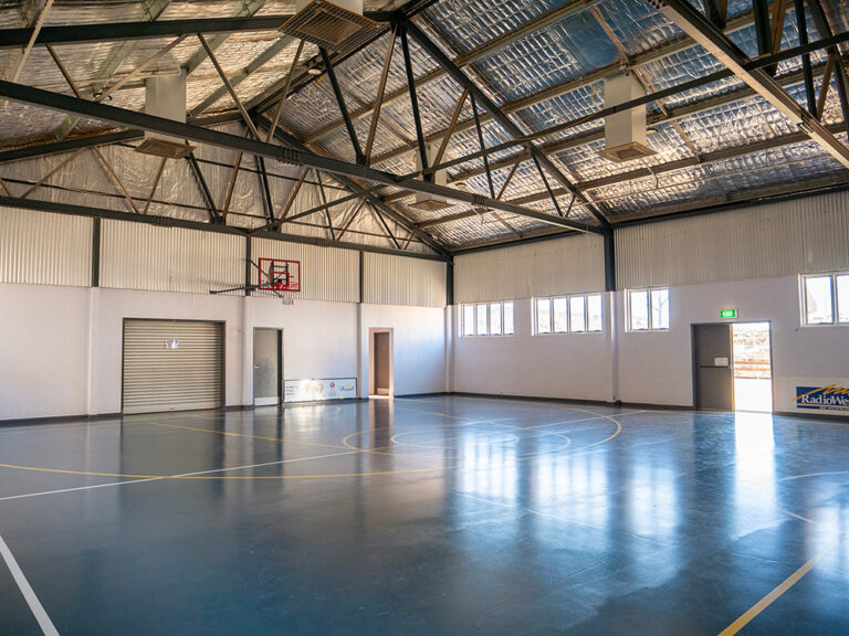 An empty indoor sports hall with a polished blue floor, basketball hoop, exposed steel roof trusses, and natural light from windows