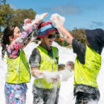 Three children play in a sea of foam outdoors on a sunny day, laughing and throwing foam at each other