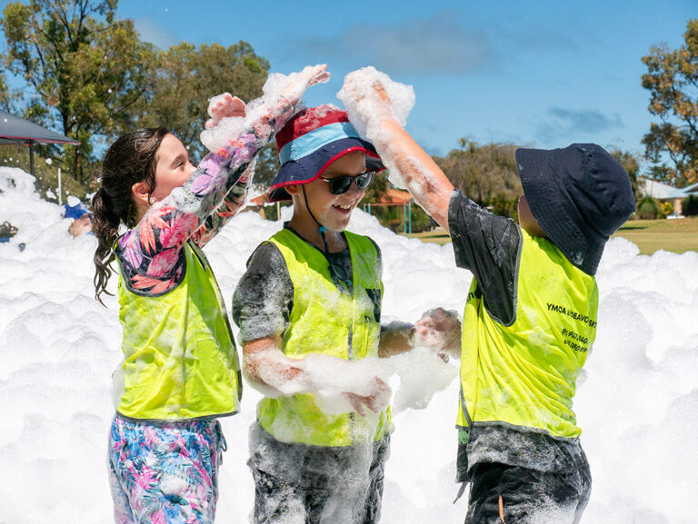 Three children play in a sea of foam outdoors on a sunny day, laughing and throwing foam at each other