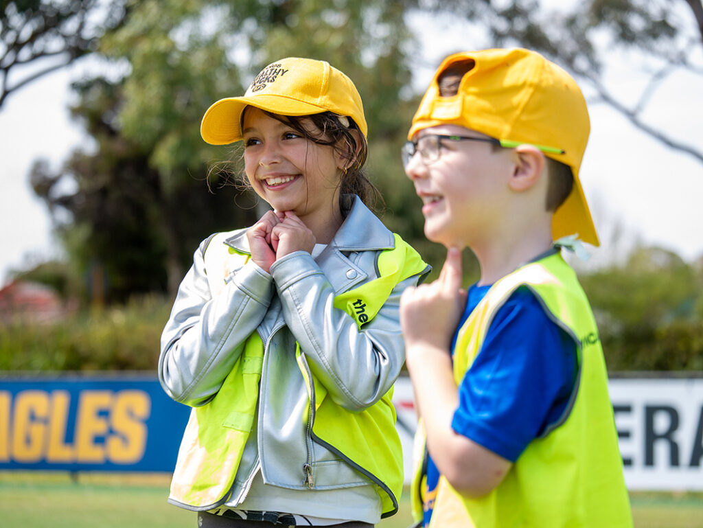 Two children wearing yellow caps and hi-vis vests smile and chat together on a sports oval