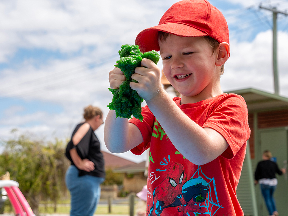 A young boy in a red cap and Spiderman t-shirt grins as he squeezes a handful of green playdough outdoors.