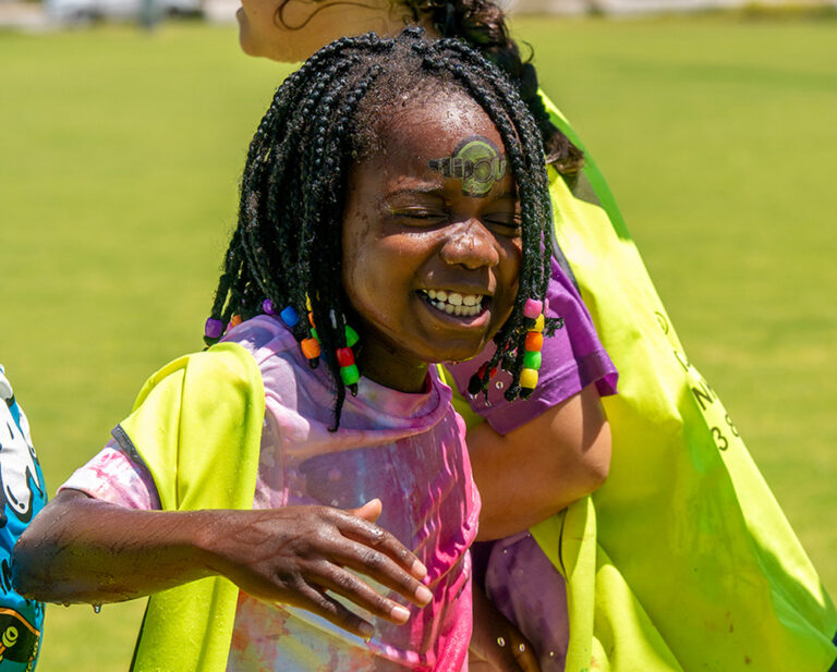 A young girl laughs with joy, face wet and decorated with a sticker, during an outdoor water play activity