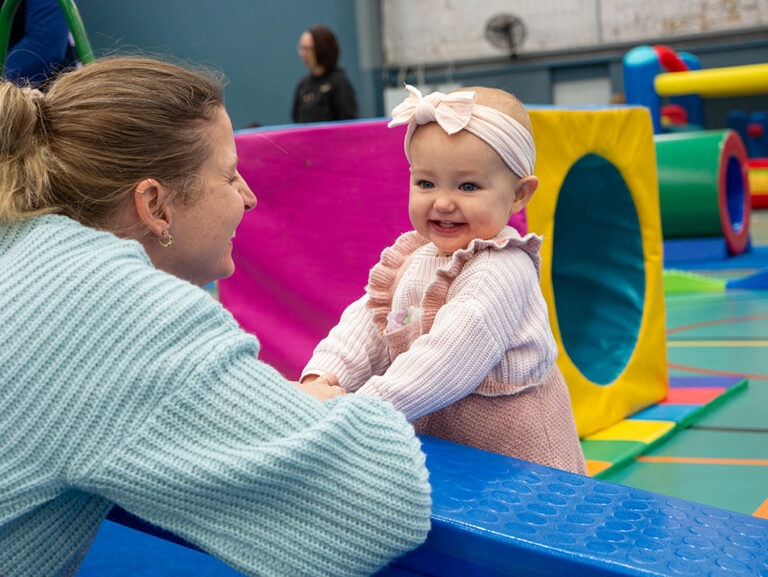 A baby in a pink knit outfit beams at her mother while sitting on soft play equipment during a parent-and-child play session