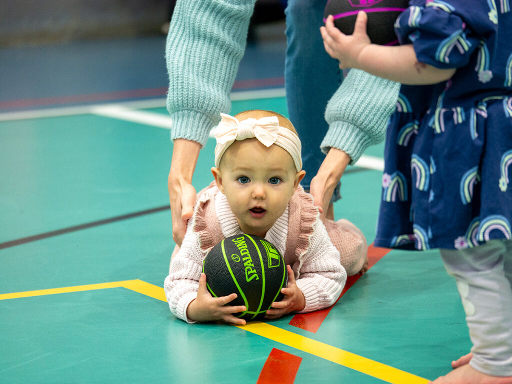 A baby in a pink outfit lies on a gym floor holding a Spalding basketball, steadied by an adult's hands, while another toddler stands nearby