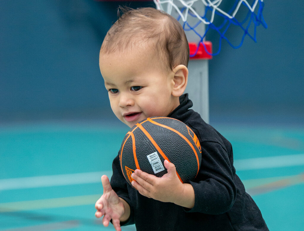 A smiling toddler in a black outfit clutches a small basketball in front of a pint-sized basketball hoop