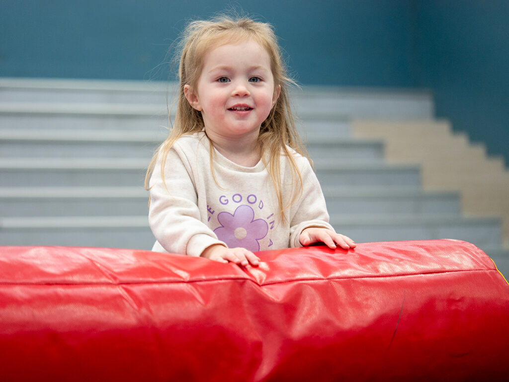 A smiling toddler in a white floral sweatshirt leans over a red foam mat during an indoor recreational play class