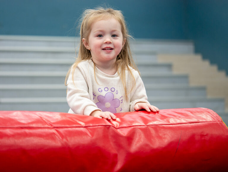 A smiling toddler in a white floral sweatshirt leans over a red foam mat during an indoor recreational play class