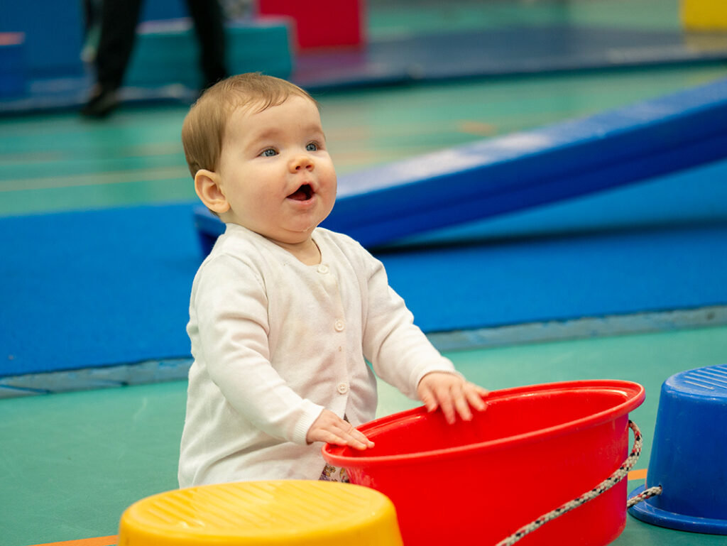A delighted baby in a white cardigan reaches toward a red plastic tub during a recreational play session on colourful soft mats.